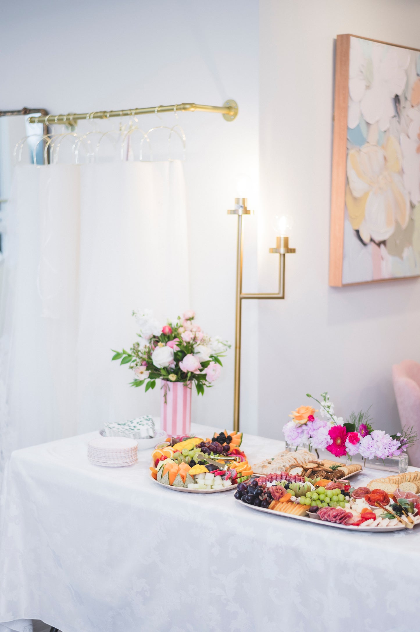 Table with fruit platters and floral arrangements in a room with white walls and a decorative painting.