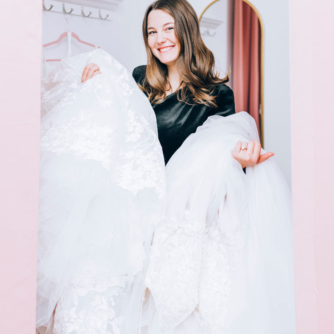 Abby holding multiple wedding dresses in a empty fitting room