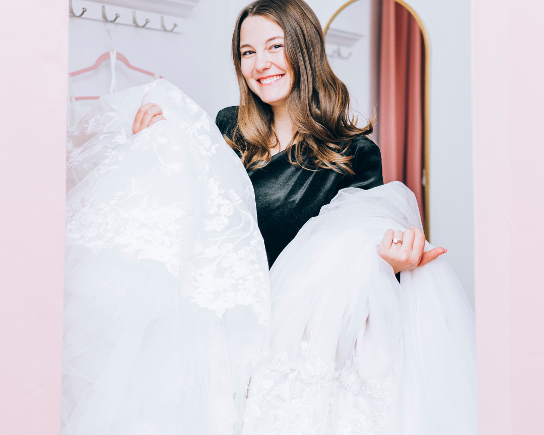 Abby holding multiple wedding dresses in a empty fitting room