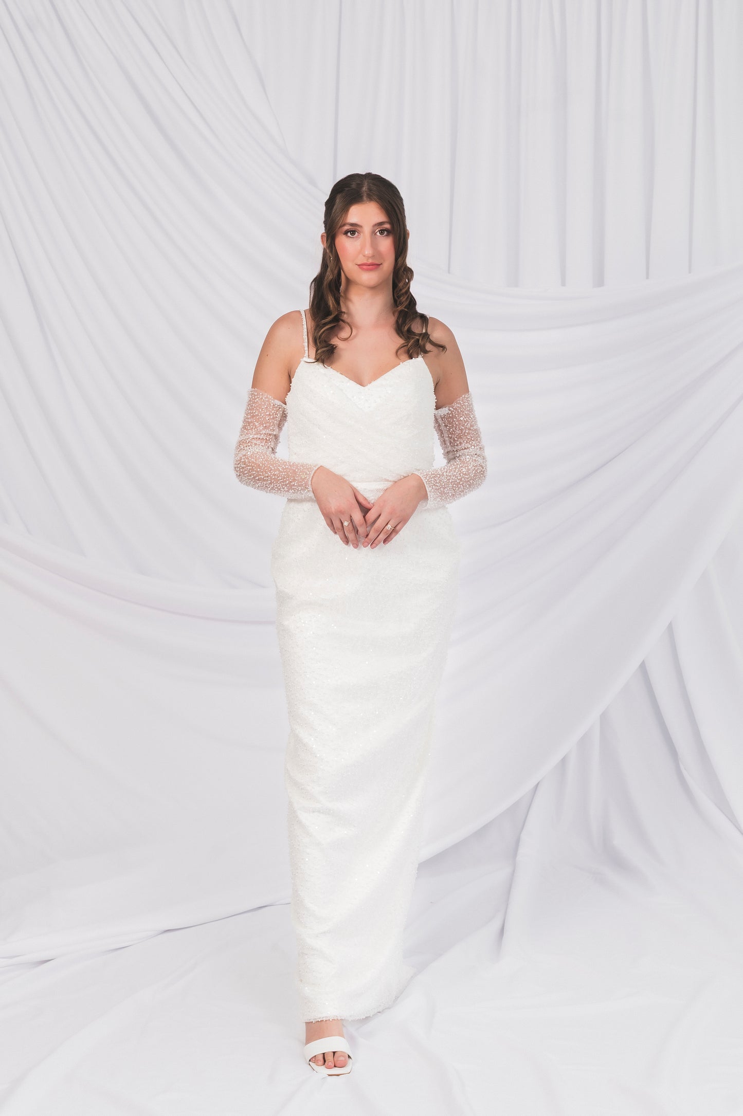 Bride wearing a white sparkly column wedding dress with sheer sparkly sleeves against a white background