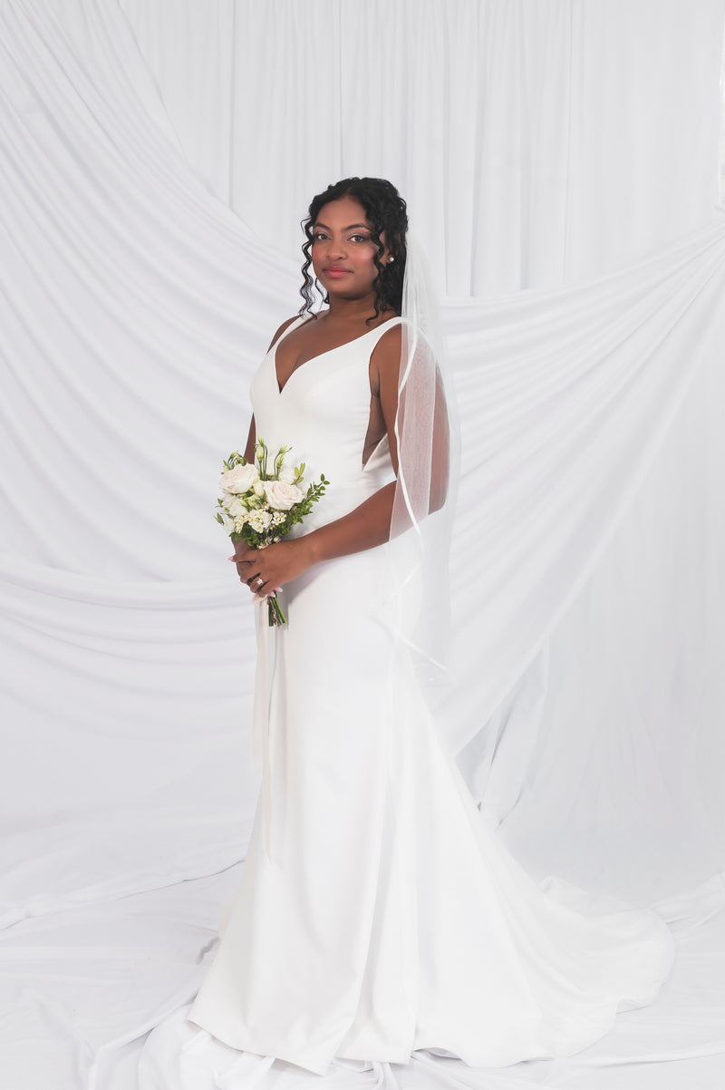 Side view of the clara gown with cut out sides, v-neck in a matte crepe fabric against a draped white wedding backdrop at Always Knew