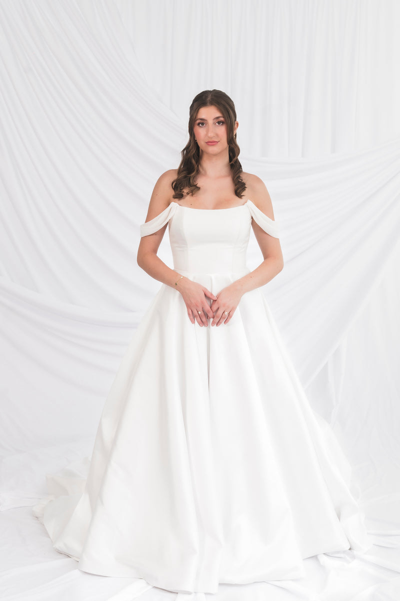 Bride in a Poppy/Clara off-shoulder wedding dress holding, Aline skirt in a silk white satin stiff mikado