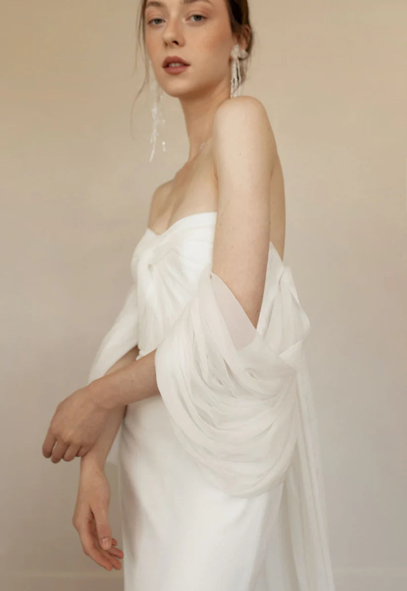 Priscilla is sleek, chic and the picture of elegance. This sheath dress is made up of crinkled chiffon and a draped bodice. Dramatic off the shoulder details and a flowy long detachable cape finish the look with perfection.