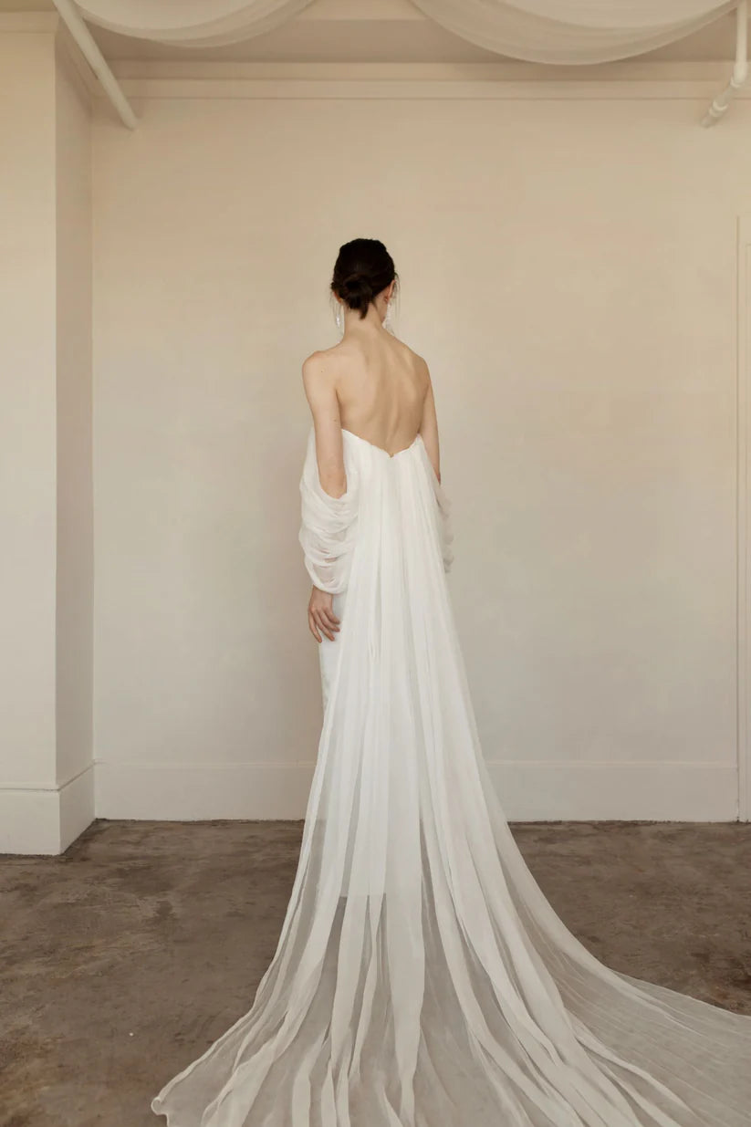 Priscilla is sleek, chic and the picture of elegance. This sheath dress is made up of crinkled chiffon and a draped bodice. Dramatic off the shoulder details and a flowy long detachable cape finish the look with perfection.