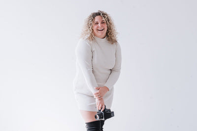 Person wearing a light gray dress and black knee-high boots holding a camera against a white background