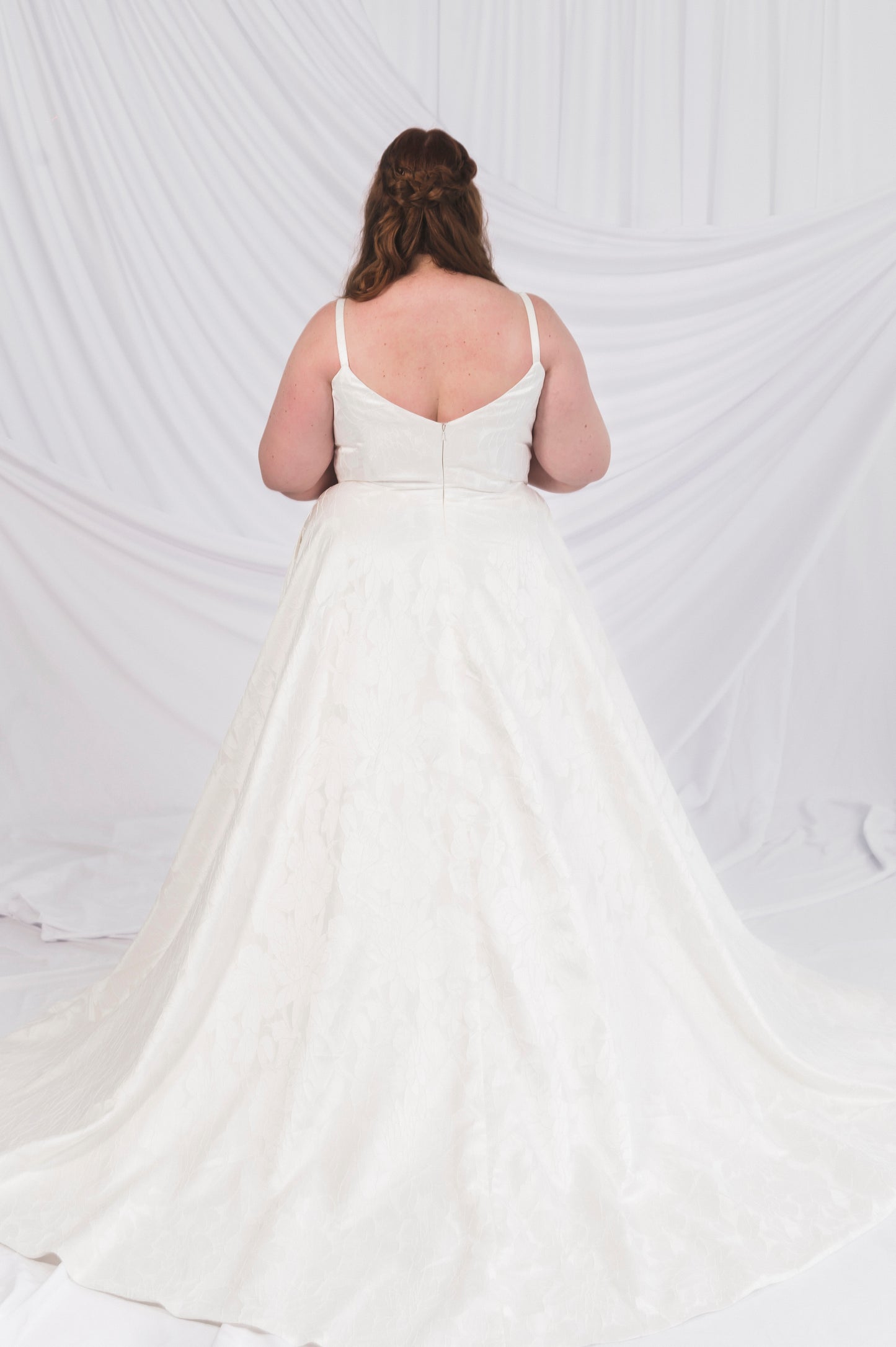 The back of a plus Size Bride wearing the Laura/Tasha, a textured simple Aline gown featuring a narrow strap, v backlin, and lighweight, but full aline skirt featuring a chapel length train. She is standing in front of a draped background in Ottawa Canada