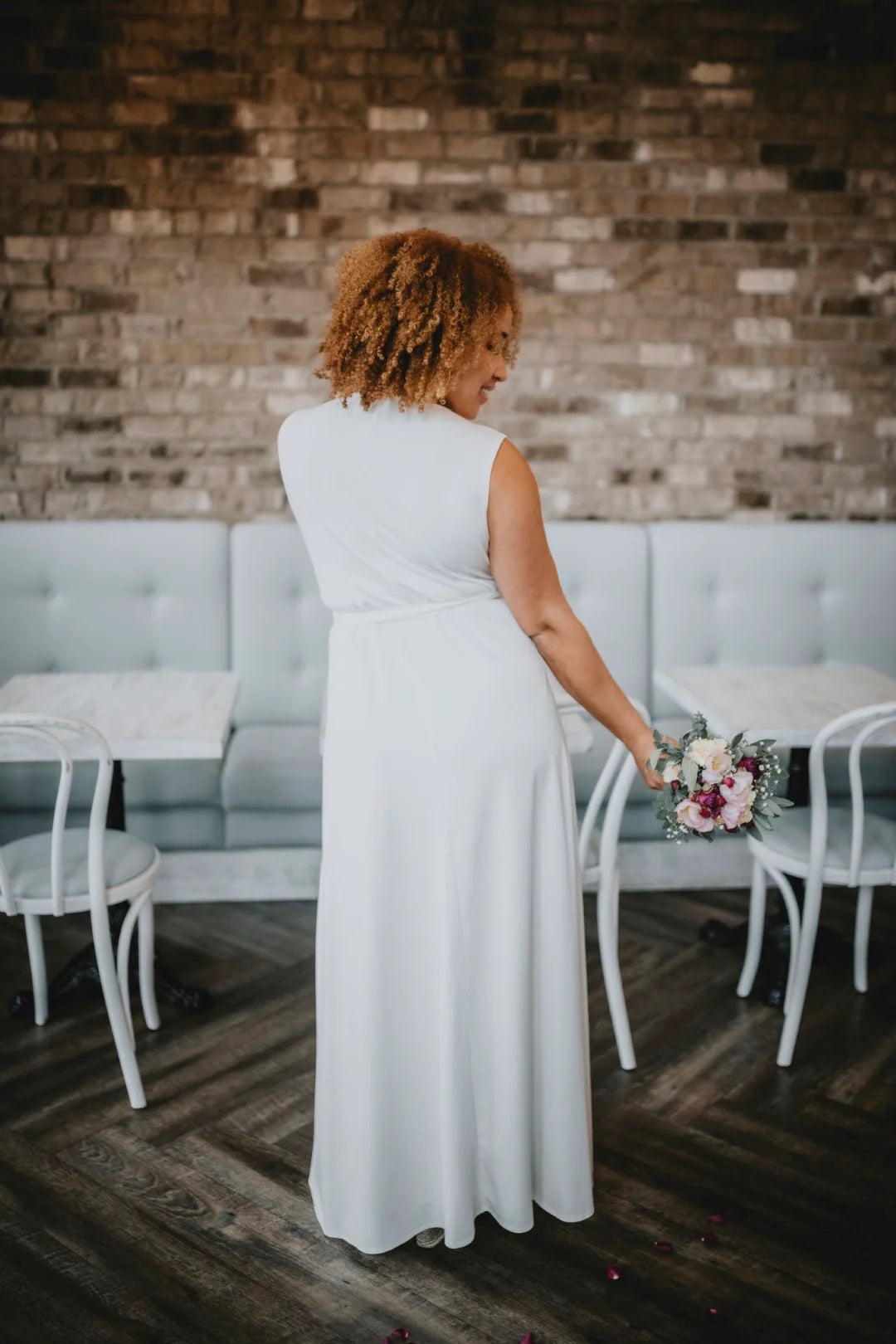 Woman in a white dress holding a bouquet in a room with a brick wall and gray furniture.