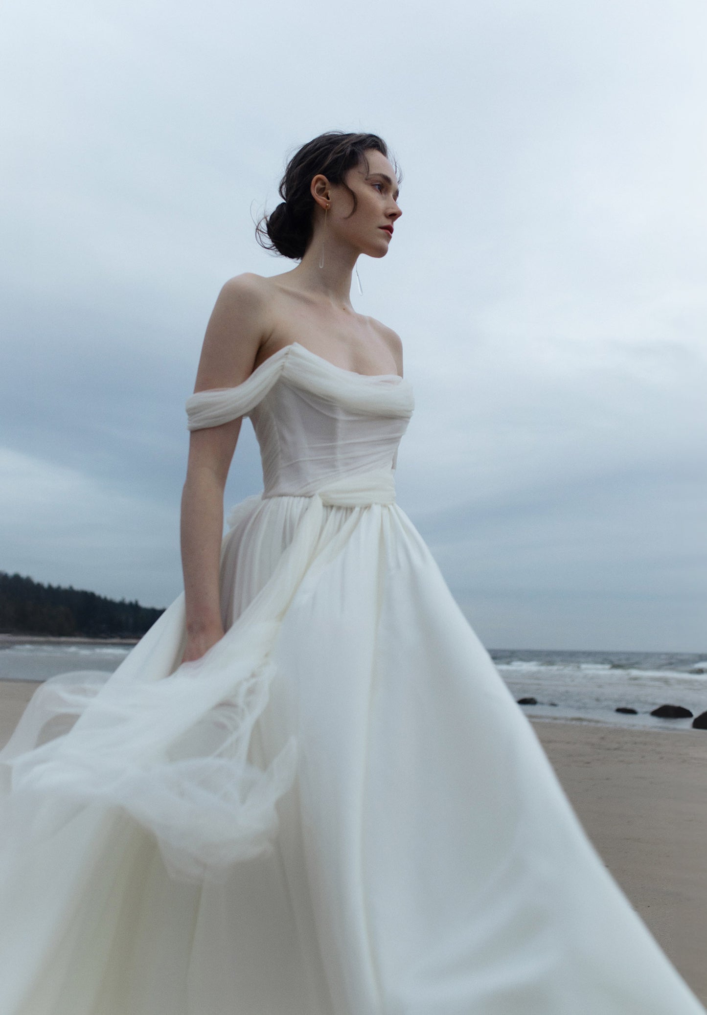 Woman in a white off-shoulder dress standing on a beach with a cloudy sky. Sora by Ouma , draped Aline wedding Dress with tied waist scarf
