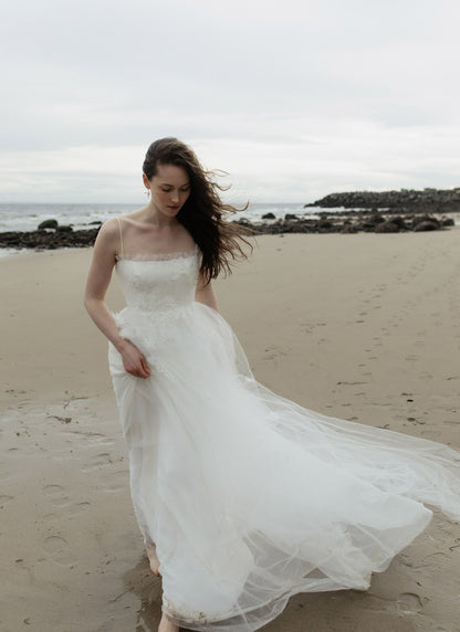 Woman in a white dress standing on a beach with ocean and sky in the background, Elise by Ouma Floral applique and square neckline