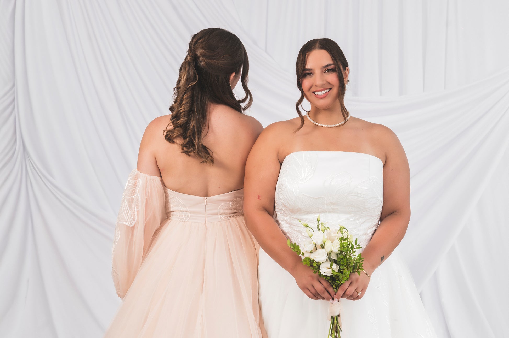 Two Bride, one in a white wedding dress and the other in a blush ballgown dress, standing against a white backgroun in  Ottawa