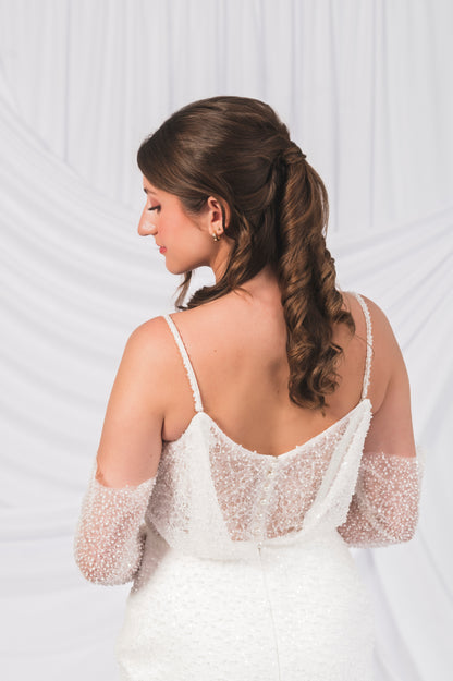 Woman in a white wedding dress with a sheer overlay and long brown hair styled in an elegant ponytail against a white background.. Sparkly V Backline with pearl buttons