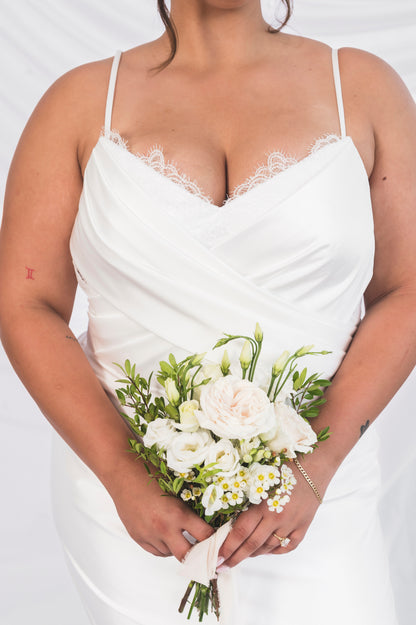 Closeup of the plus size bride italian satin bodice with a draped v neck bodice and peakaboo eyelash french lace, holding a mini bouquet from Goldie Florals