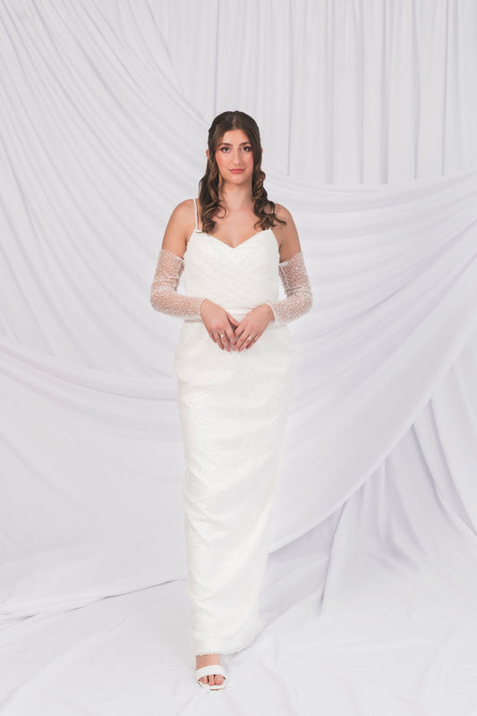 Bride wearing a white sparkly column wedding dress with sheer sparkly sleeves against a white background