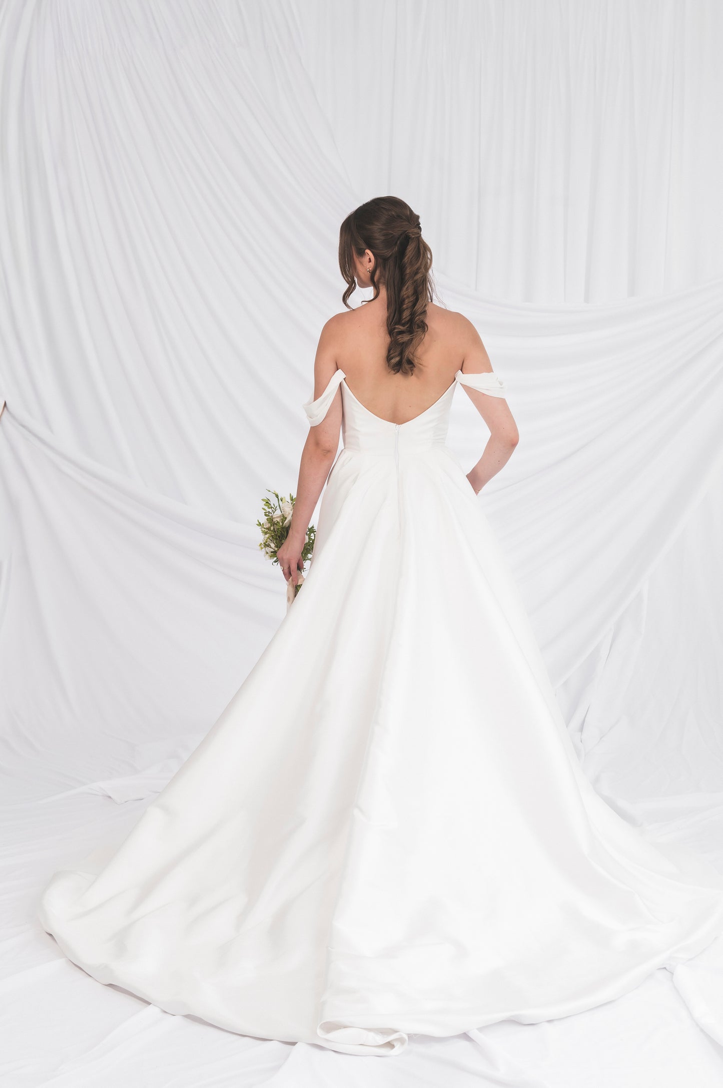 Bride in a Poppy/Clara off-shoulder wedding dress holding a bouquet against a white back ground, showing the back v scooped mikado with a dramatic a-line train in Ottawa, Always Knew Boutique