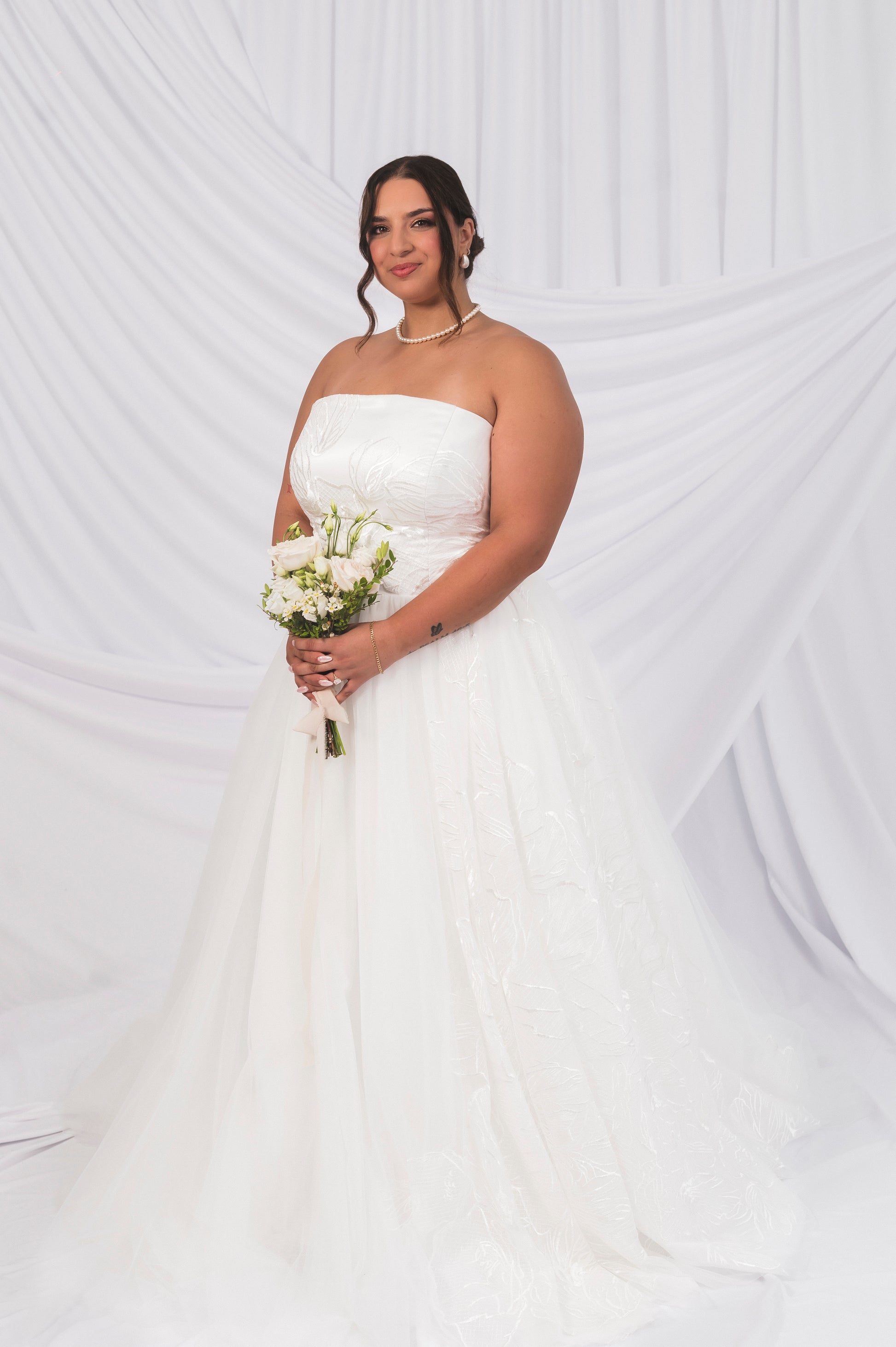 Plus Size Bride Yas wearing a straight across neckline tulle ballgown holding a mini white floral bouquets
