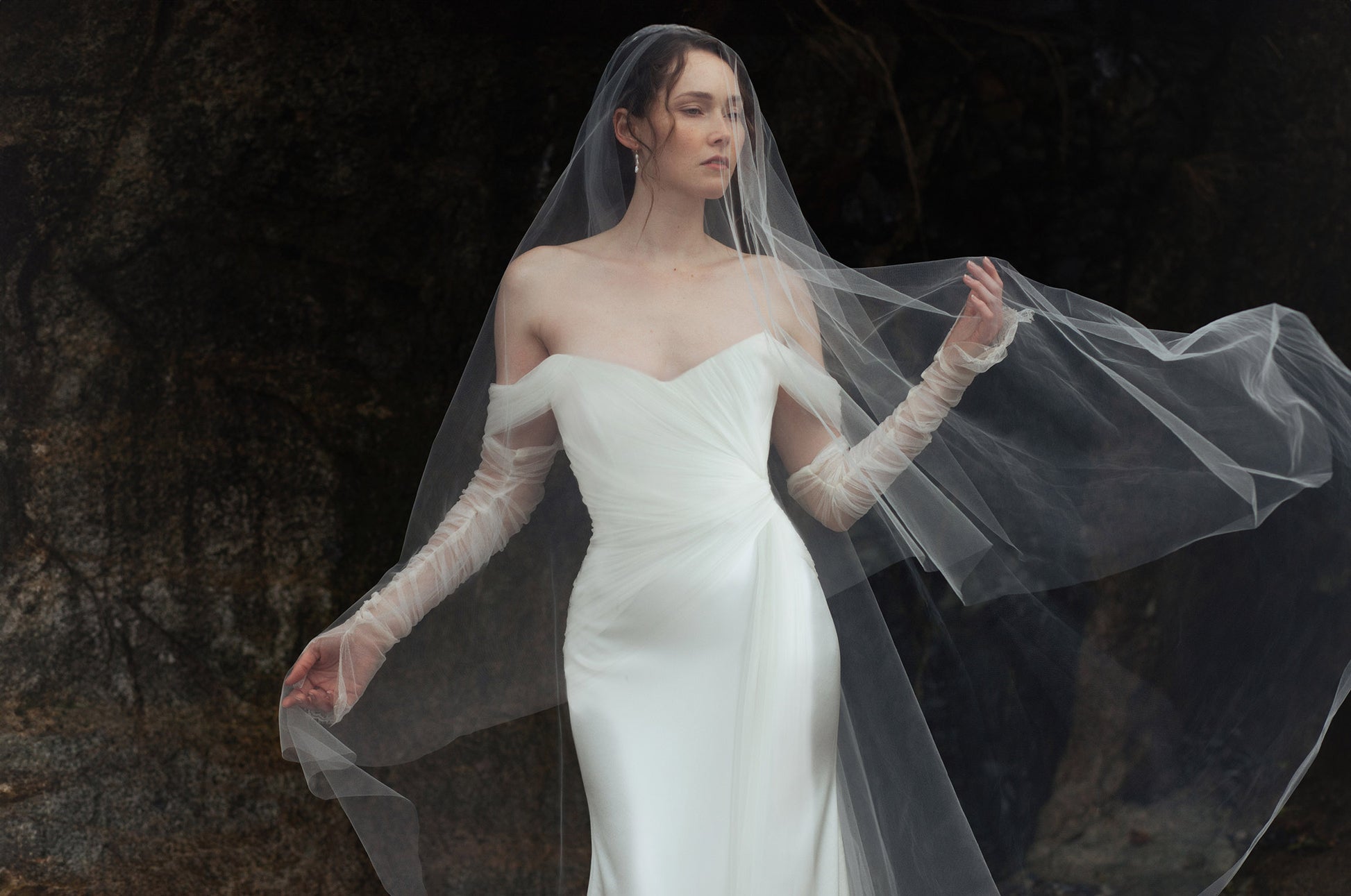 Bride in a white wedding dress, Elysian by Ouma, with  the matching veil against a dark background. Featuring the off the shoulder soft sweatheart neckline and fitted satin silk bodice 
