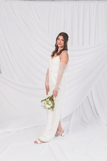Ariel/Tessa wedding dress column shift dress with the removable gloves. Bride is holding a mini bouquet from Goldie Florals and shows off the back slit of the dress, with no train, against a draped backdrop