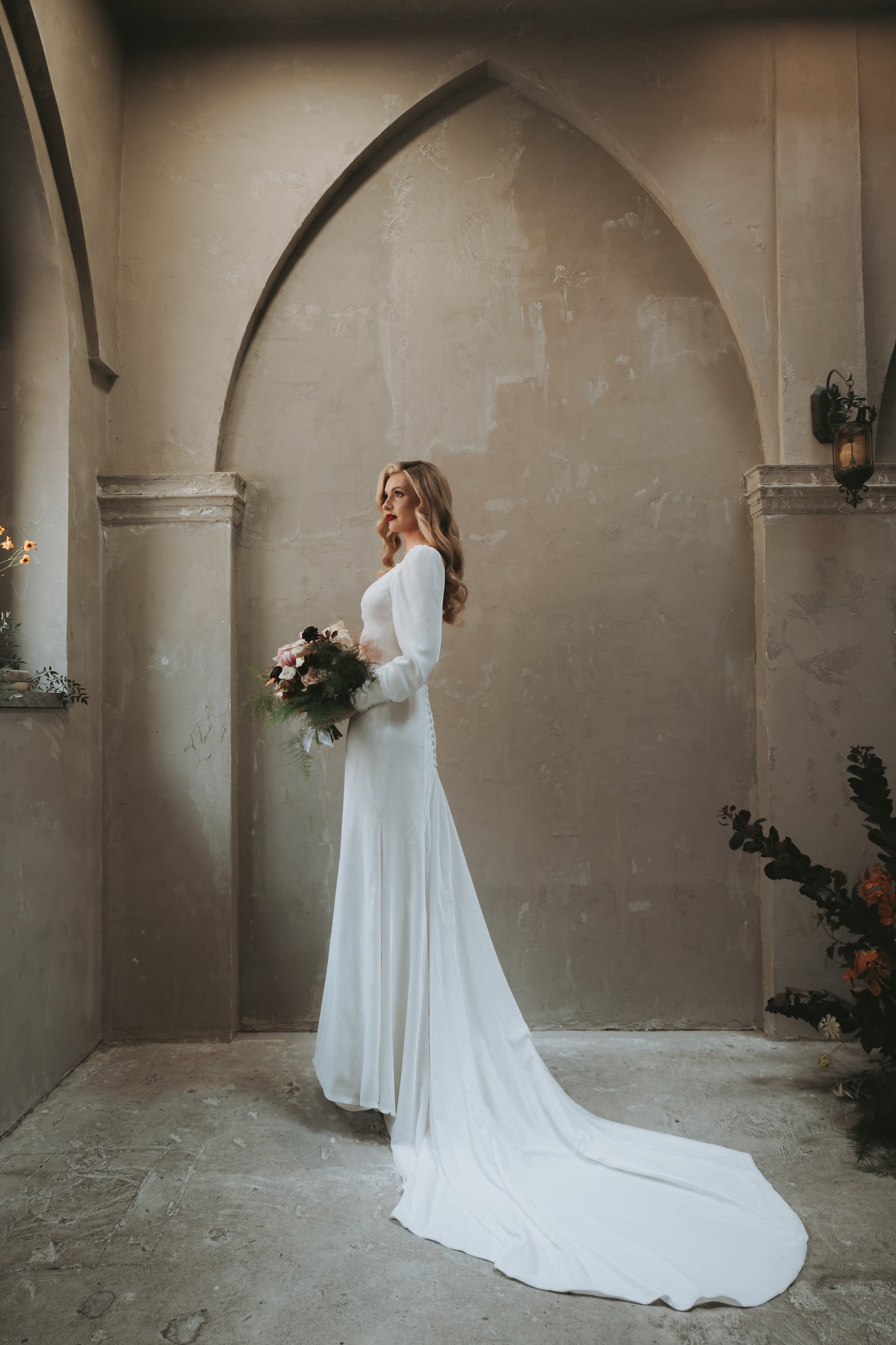 Woman in a white long sleeve modest wedding dress holding a bouquet in an elegant interior setting. Velvet material and custom bishop sleeves