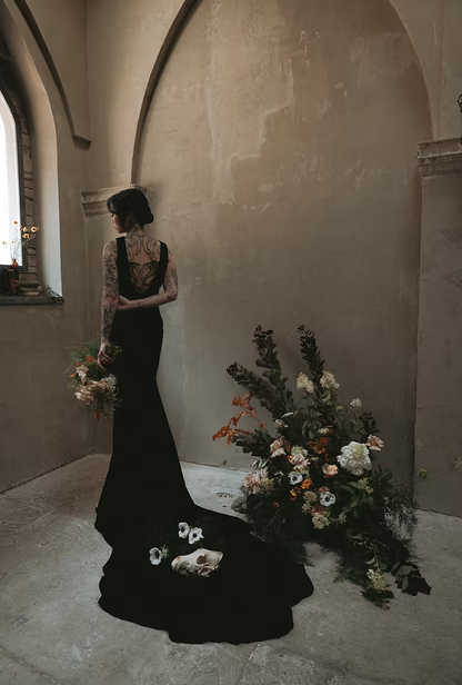 Woman in a black wedding dress. showing off the low back of the velvet gown for a gothic wedding portrait, standing in a room with floral arrangements.