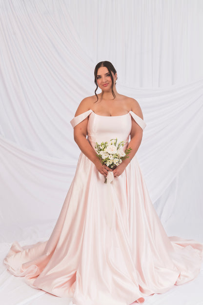 Front View Bride wearing the Poppy/Laura Blush Satin Gown, scooped soft cat-eye neckline with off the shoulder satin drapes, holding a bouquet against a soft draped backdrop in Ottawa Canada