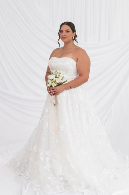 Front View of Plus Size Bride wearing classic lace option of the strapless poppy/clara dress straight neckline and floral pattern, holding a bouquet