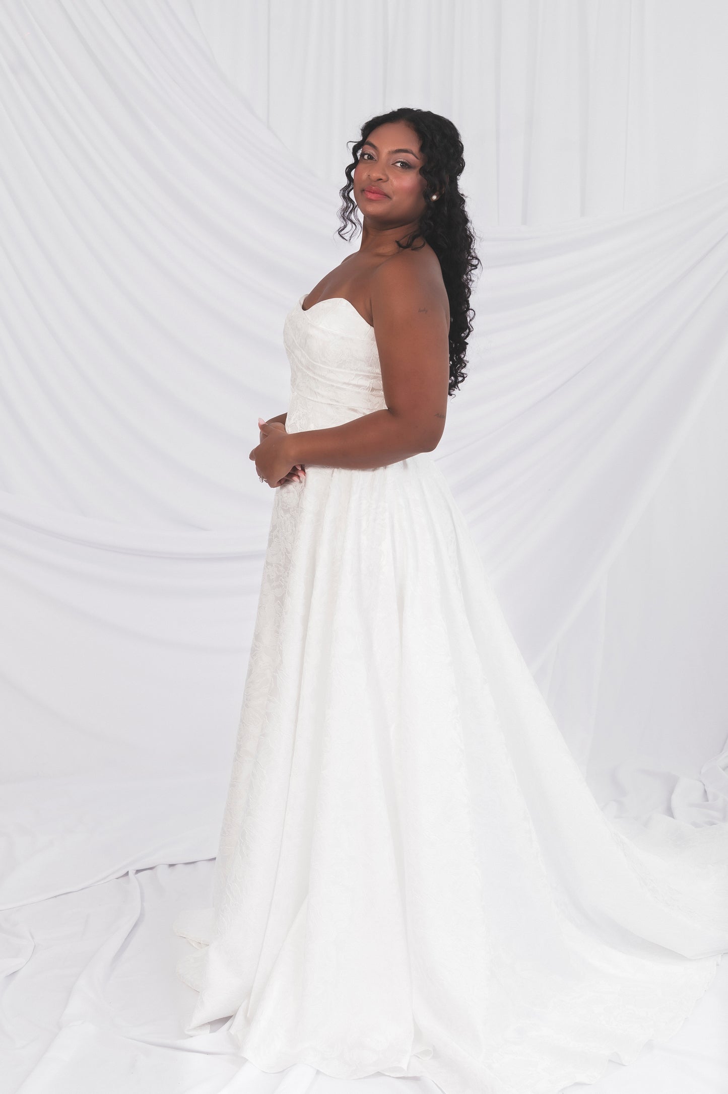 Side view of the klein basque wedding dress, soft sweetheart neckline and long train. Worn on a size 10 model in front of a draped backdrop in Ottawa