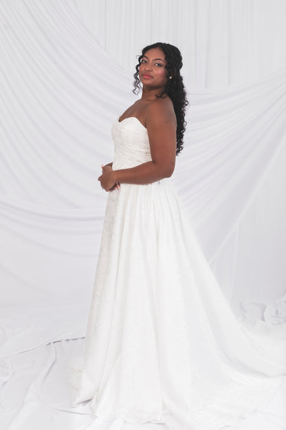 Side view of the klein basque wedding dress, soft sweetheart neckline and long train. Worn on a size 10 model in front of a draped backdrop in Ottawa