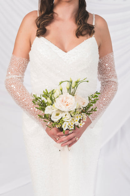 Bride holding a bouquet of flowers from Goldie Florals, with sparkly gloves on a light background. V neck with draped bodice and narrow straps , beaded