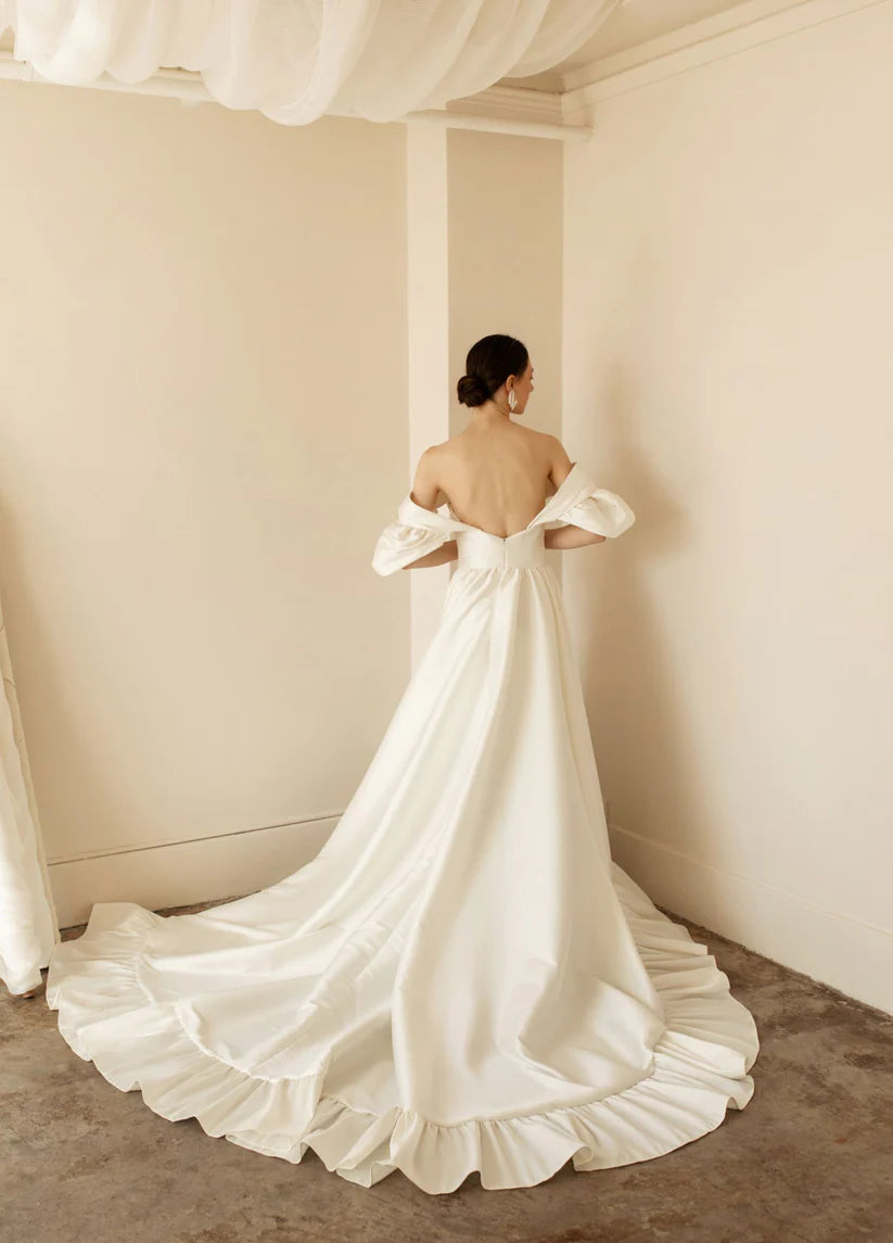 Hazel is a statement dress. Made of luxurious Mikado, this a-line piece with a peek-a-boo twisted bodice is a show stopper. A gathered skirt with a ruffled edge and a subtle leg slit create the dream gown.