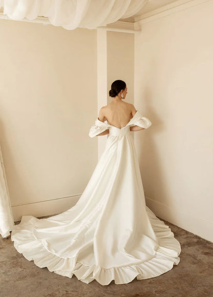 Hazel is a statement dress. Made of luxurious Mikado, this a-line piece with a peek-a-boo twisted bodice is a show stopper. A gathered skirt with a ruffled edge and a subtle leg slit create the dream gown.