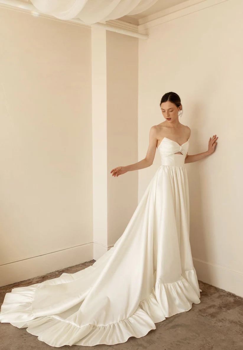 Hazel is a statement dress. Made of luxurious Mikado, this a-line piece with a peek-a-boo twisted bodice is a show stopper. A gathered skirt with a ruffled edge and a subtle leg slit create the dream gown.