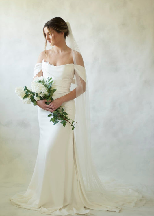 Juniper is a pearl crepe gown featuring a draped bodice paired with a fit and flare skirt featuring a slit. Embroidery lace appliques with pearl beading scatter through out the gown, and pearl buttons detail the zipper line