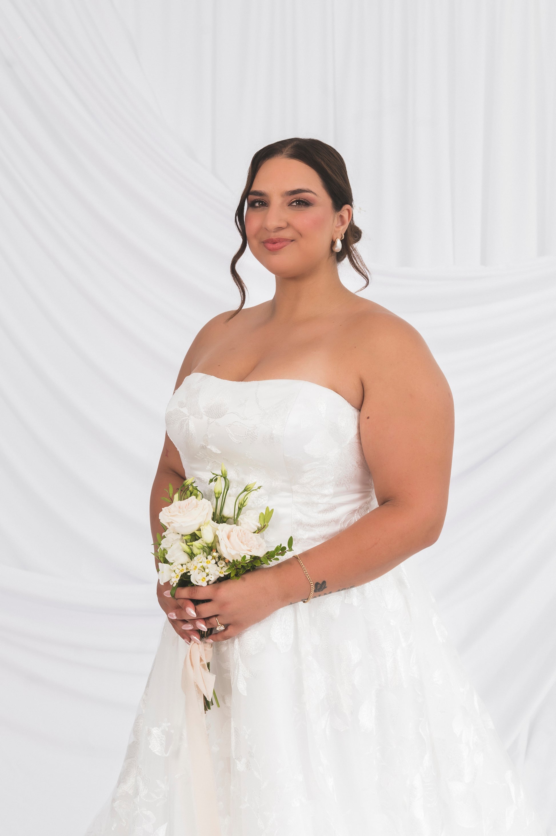 closeup of Plus Size Bride wearing classic lace option of the strapless poppy/clara dress straight neckline and floral pattern, holding a bouquet