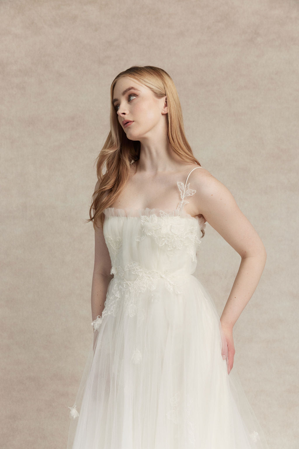 Woman wearing a white thin strap square neckline wedding dress, Elise by Ouma against a beige background. Closeup of bodice with sporadic lightweight floral appliquee
