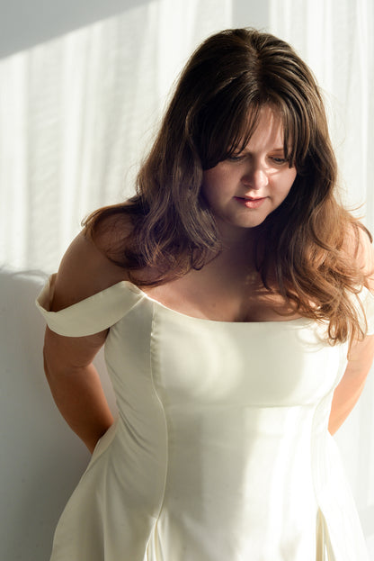 Closeup of designer Kathryn bass wearing a the poppy/laura bridal gown with a structured bodice and pleated skirt, standing in a room with draped curtains. Shown here with the draped sleeve