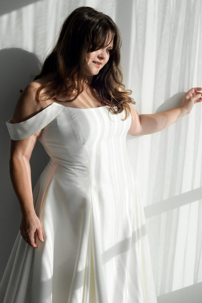 Designer Kathryn bass wearing a the poppy/laura bridal gown with a structured bodice and pleated skirt, standing in a room with draped curtains. Shown here with the draped sleeve
