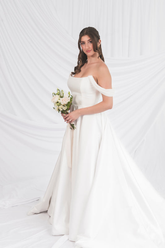 Bride in a Poppy/Clara off-shoulder wedding dress holding a bouquet against a white background in Ottawa, Always Knew Boutique