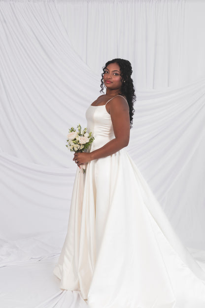 Side view of the bride wearing the poppy/laura, the structured bodice with a pleated skirt and a satin narrow strap against a draped background in Ottawa at Always Knew.