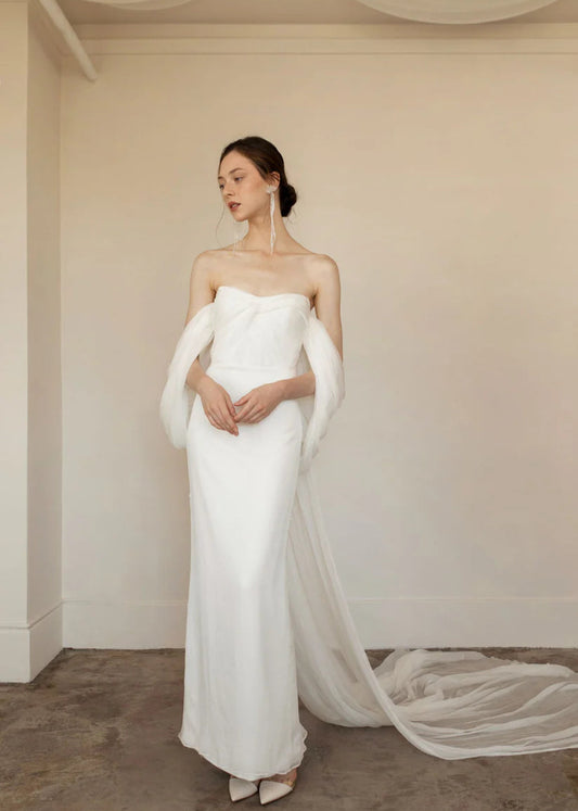 Priscilla is sleek, chic and the picture of elegance. This sheath dress is made up of crinkled chiffon and a draped bodice. Dramatic off the shoulder details and a flowy long detachable cape finish the look with perfection.