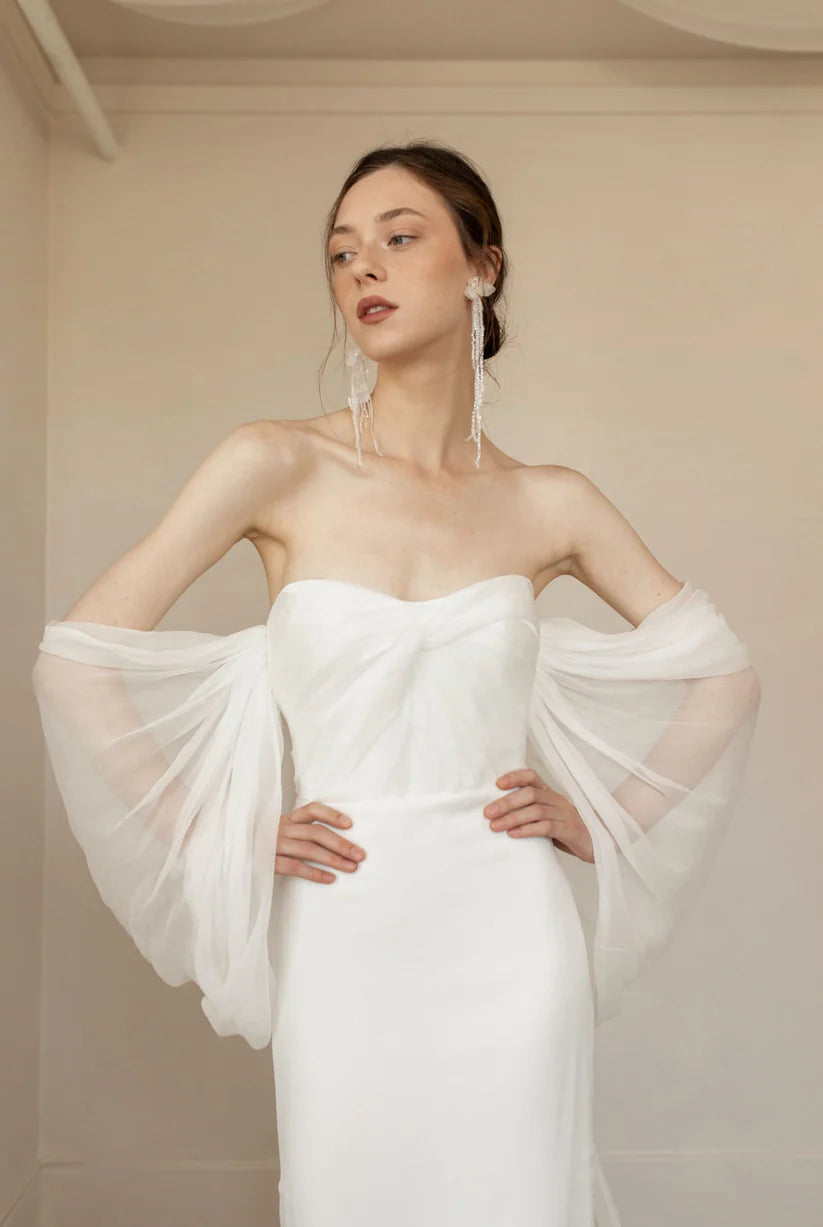 Priscilla is sleek, chic and the picture of elegance. This sheath dress is made up of crinkled chiffon and a draped bodice. Dramatic off the shoulder details and a flowy long detachable cape finish the look with perfection.