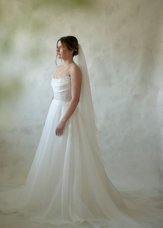 A satin organza gown featuring a full skirt, flowing train and semi sheer corset bodice with a button closure. The gown is accented by bust draping and pearl beaded appliques scattering throughout the gown