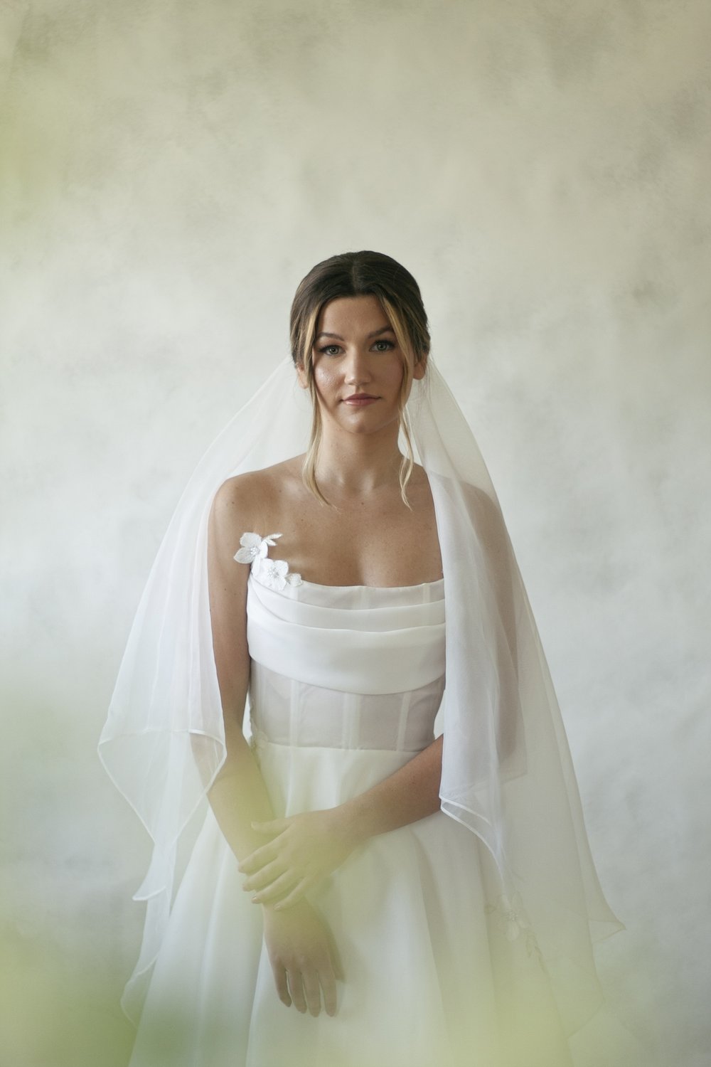 A satin organza gown featuring a full skirt, flowing train and semi sheer corset bodice with a button closure. The gown is accented by bust draping and pearl beaded appliques scattering throughout the gown
