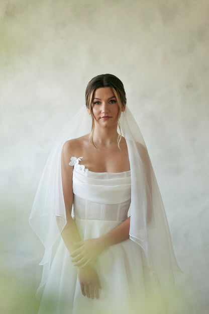 A satin organza gown featuring a full skirt, flowing train and semi sheer corset bodice with a button closure. The gown is accented by bust draping and pearl beaded appliques scattering throughout the gown