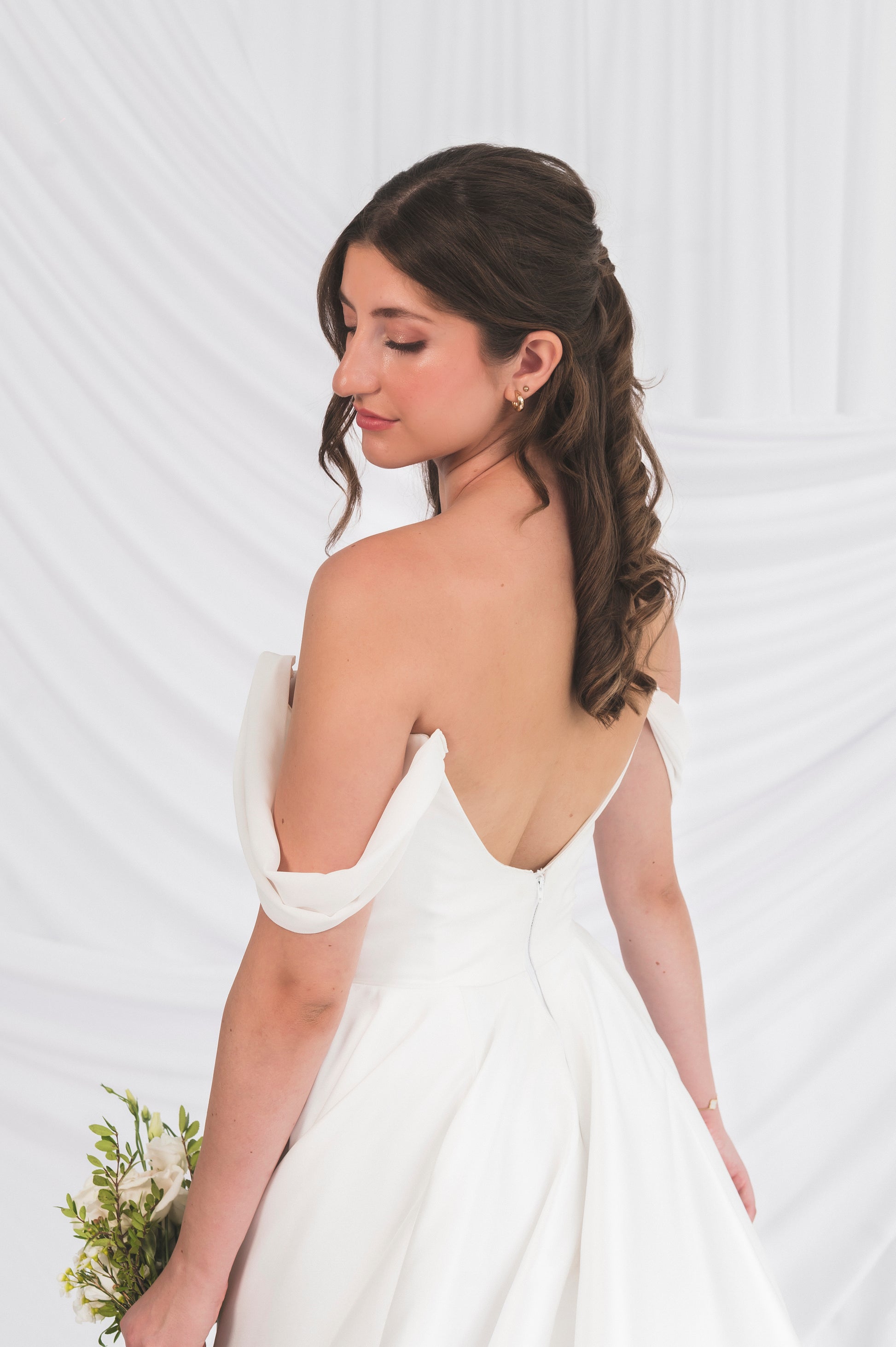 Closeup of a Bride in front of a draped white background at Always Knew Bridal boutique in ottawwa. She is showing off the scooped low back of the Poppy/Clara dress with the chiffon off the shoulder add on straps