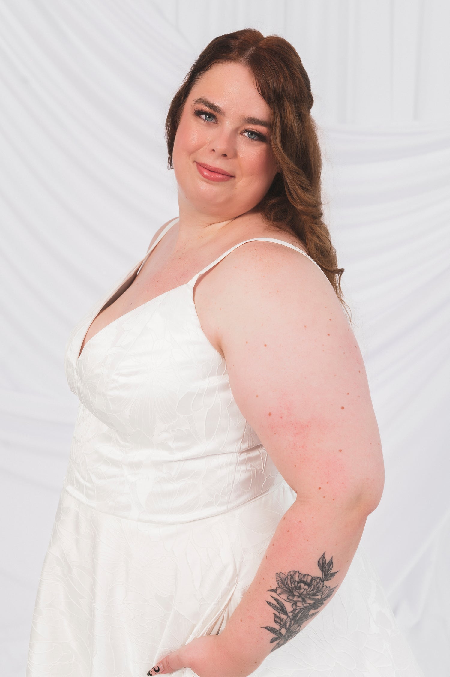 Closeup of the side of Plus Size Bride wearing the Laura/Tasha, a textured simple Aline gown featuring a narrow strap, v neck bodice, and lighweight, but full aline skirt. She is standing in front of a draped background in Ottawa Canada