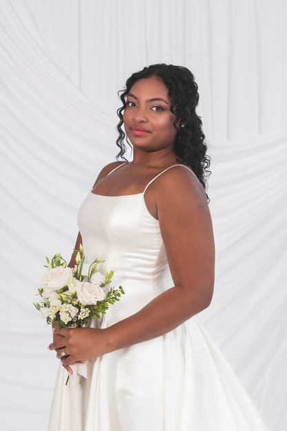 Closeup of the side view of the bride wearing the poppy/laura, the structured bodice with a pleated skirt and a satin narrow strap against a draped background in Ottawa at Always Knew.