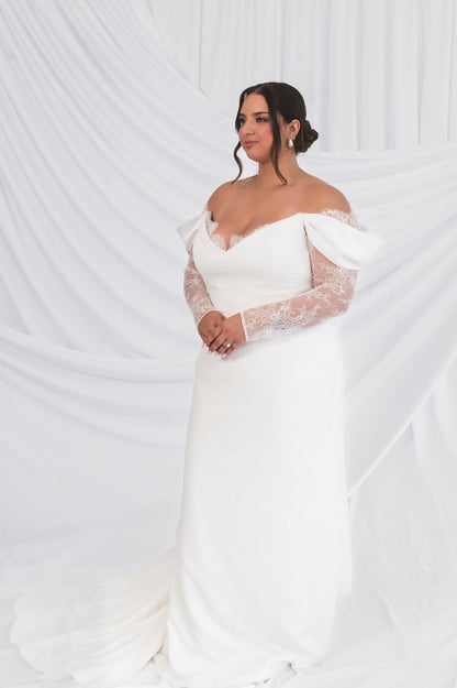 Bride wearing a white off-shoulder wedding dress with sheer sleeves against a white background, the Jane/Vicki including eyelash lace