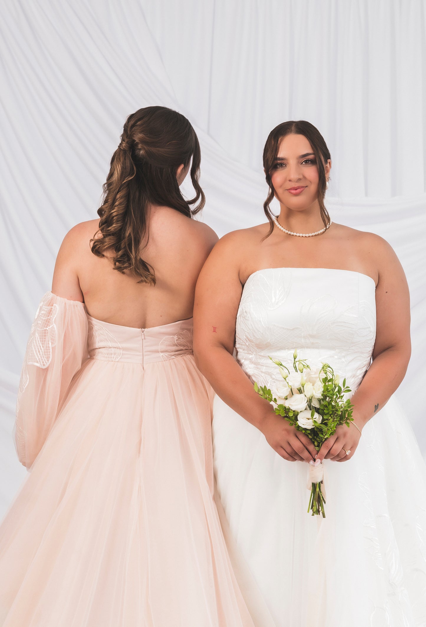 Two Brides wearing the Briar Ball Gown. One bride is showing off the back of the blush briar gown with oversized detachable sleeve. Our Plus size Bride is wearing the Briar strapless tulee gown in the white