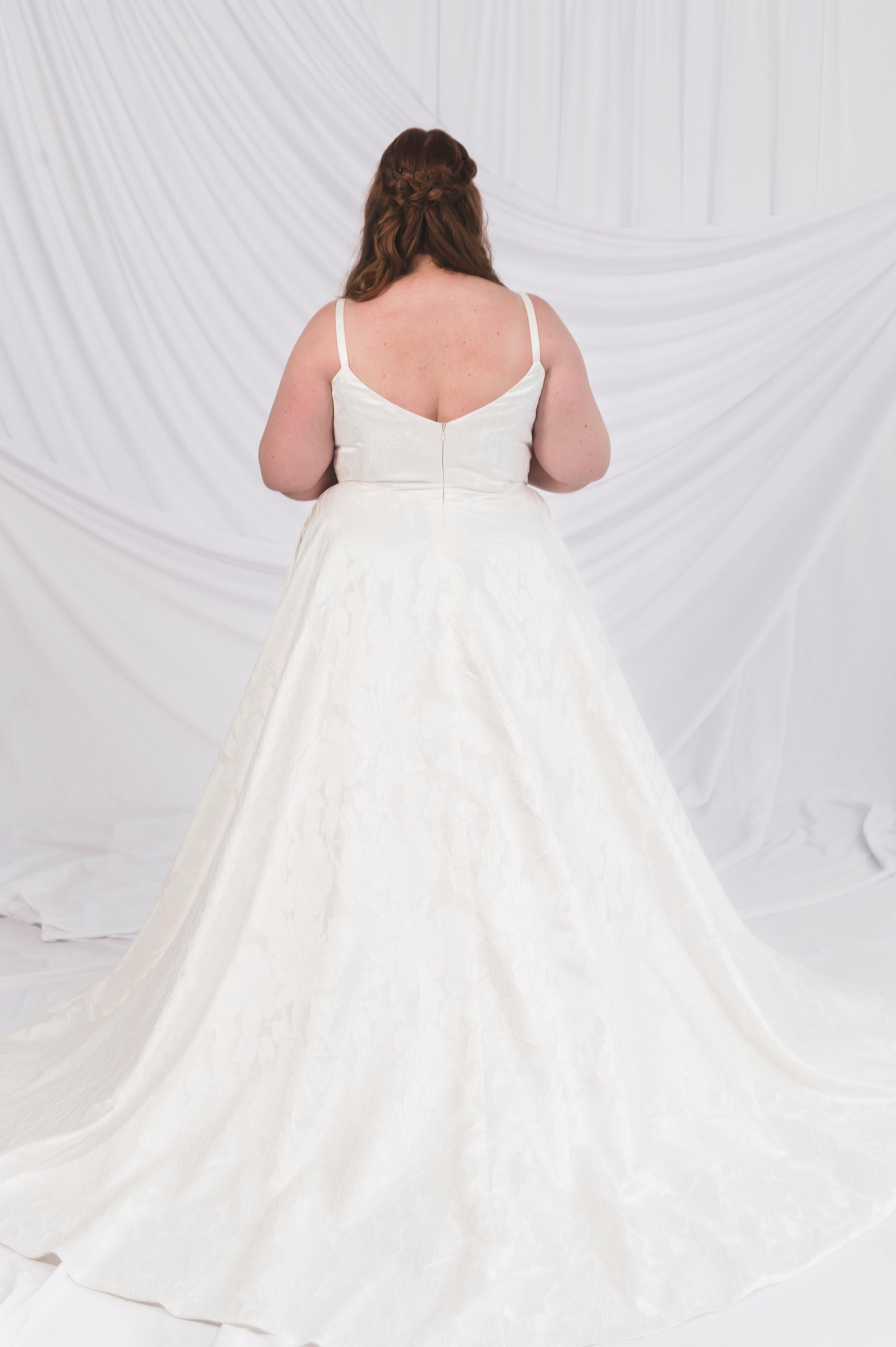 The back of a plus Size Bride wearing the Laura/Tasha, a textured simple Aline gown featuring a narrow strap, v backlin, and lighweight, but full aline skirt featuring a chapel length train. She is standing in front of a draped background in Ottawa Canada