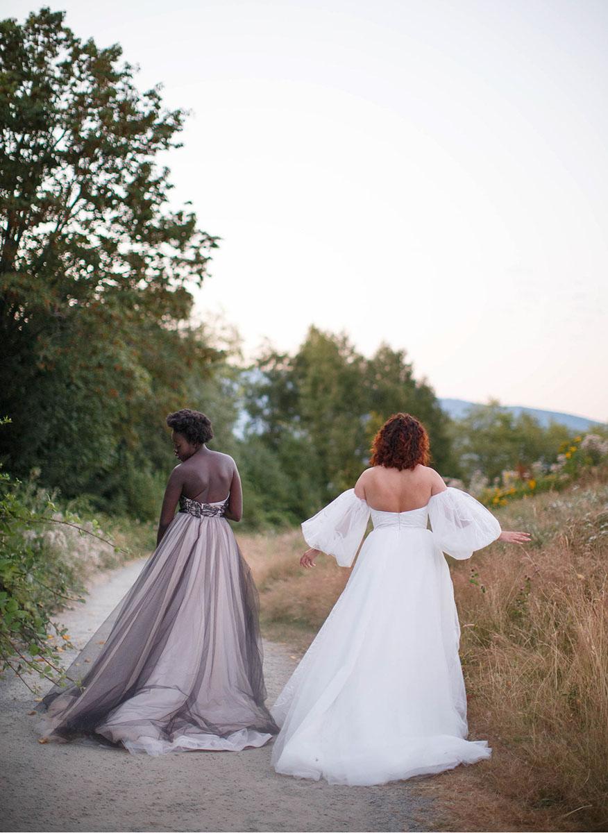 Two Brides in the straight neckline with a large tulle skirt,  walking away on a path with trees and mountains in the background.